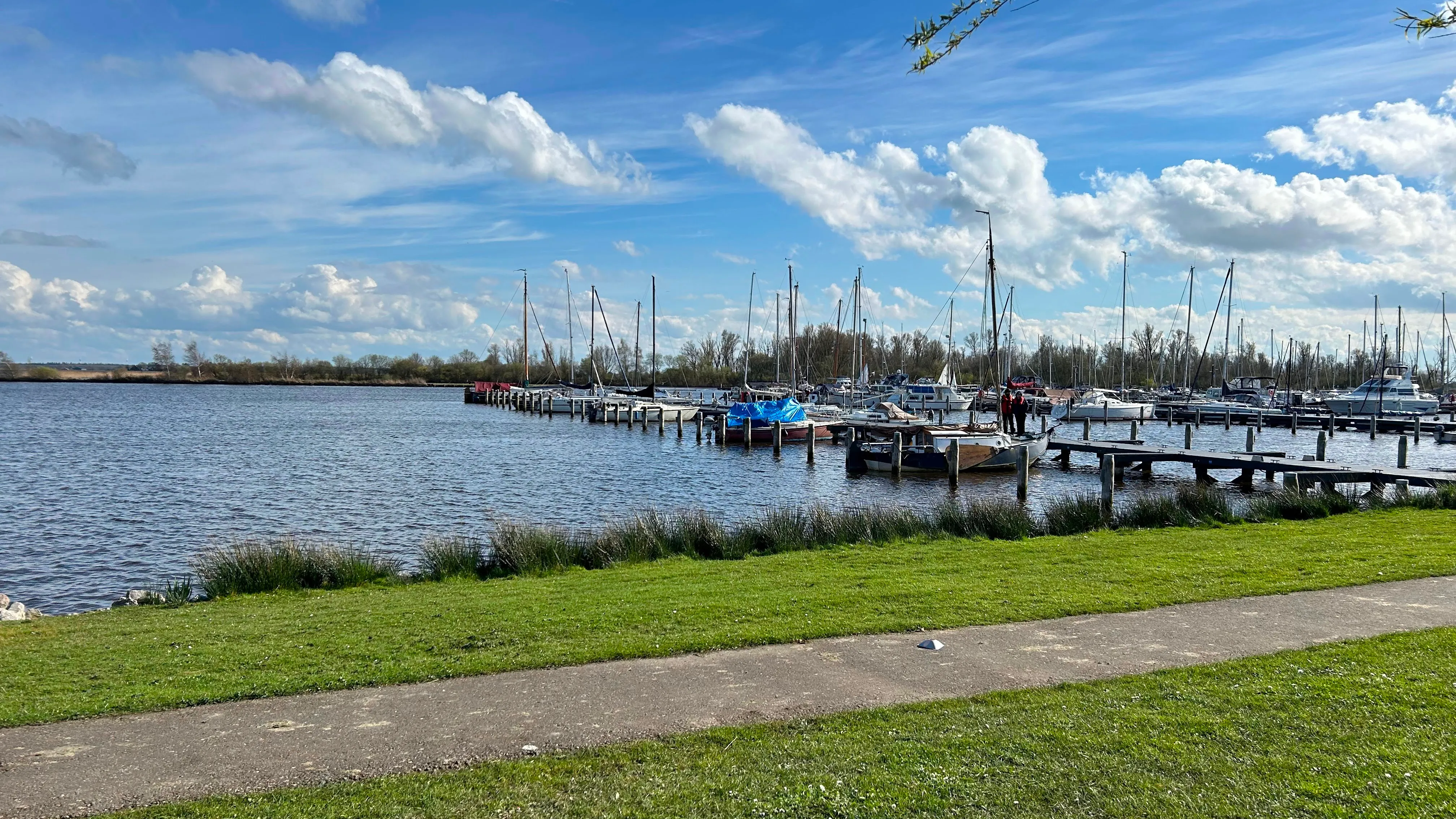 Koudum harbor under broken clouds — sailboats moored along the piers, grass in the foreground.
